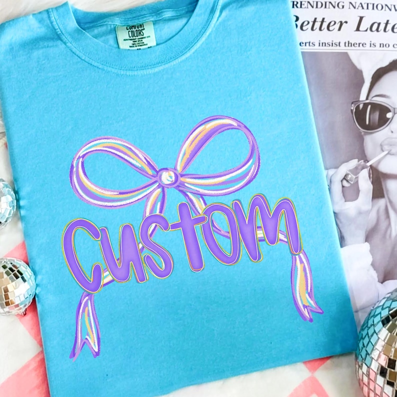 May include: A light blue t-shirt featuring a large purple bow and the word "Custom" in a purple script font. The shirt has a Comfort Colors label. Two disco balls are visible in the background.