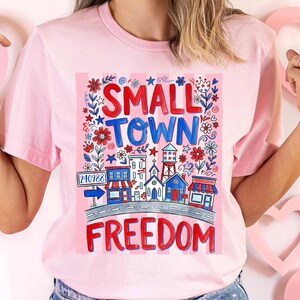 May include: Pink t-shirt with a colorful graphic design. The design features the words "SMALL TOWN FREEDOM" in red and blue, along with illustrations of buildings, flowers, and stars. The shirt has short sleeves and a crew neck.