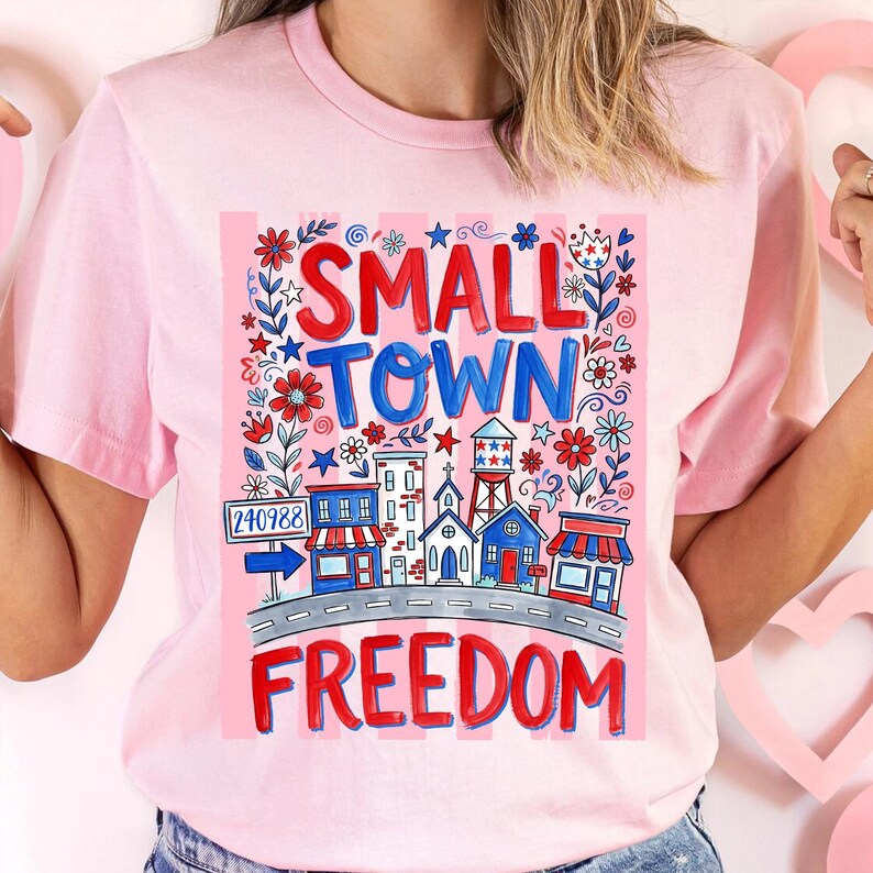 May include: Pink t-shirt with a colorful graphic design. The design features the words "SMALL TOWN FREEDOM" in red and blue, along with illustrations of buildings, flowers, and stars. The shirt has short sleeves and a crew neck.