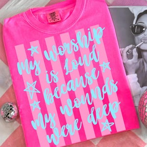 May include: A bright pink t-shirt with vertical light pink stripes and the text "My worship is loud because my wounds were deep" in light blue script. The shirt has a crew neck and short sleeves. The brand label "Comfort Colors" is visible.