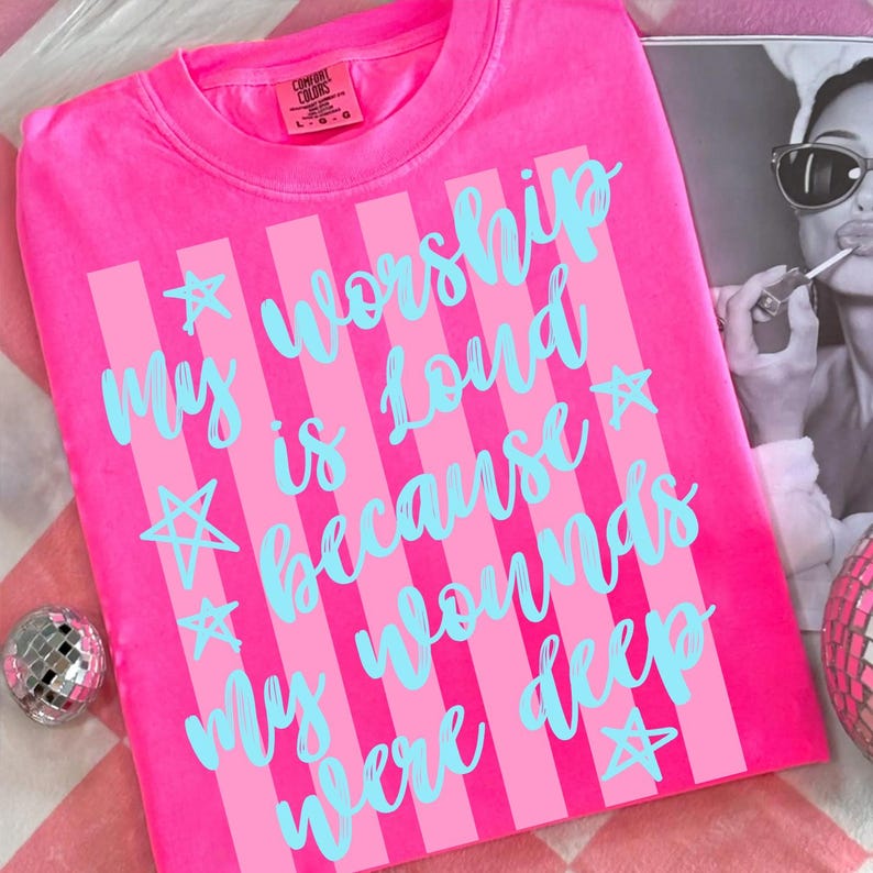 May include: A bright pink t-shirt with vertical light pink stripes and the text "My worship is loud because my wounds were deep" in light blue script. The shirt has a crew neck and short sleeves. The brand label "Comfort Colors" is visible.