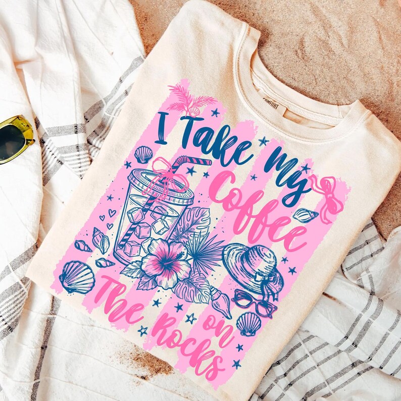 May include: Cream-colored sweatshirt with a graphic that reads "I Take My Coffee on The Rocks." The design features a tropical theme with a drink, flowers, seashells, and a hat in pink and blue. The shirt is folded on a white and gray striped towel.