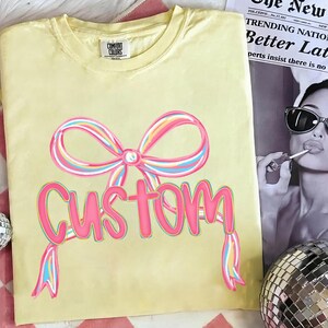 May include: A pale yellow Comfort Colors t-shirt with a large, multi-coloured bow design above the word "Custom" in pink, with a matching ribbon. The shirt is displayed on a white surface with a newspaper and disco ball in the background.