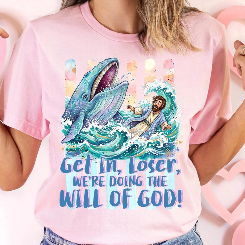 May include: Pink t-shirt featuring a whimsical illustration of a blue whale with an open mouth, a man in a robe, and ocean waves. The text "Get In, Loser, We're Doing The Will Of God!" is printed in blue.