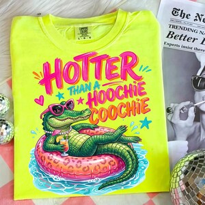 K&ouml;nnte beinhalten: Ein neongelbes T-Shirt mit dem Schriftzug "Hotter Than a Hoochie Coochie" in Pink und Orange. Die Grafik zeigt ein gr&uuml;nes Alligator mit Sonnenbrille und Halskette, der auf einem aufblasbaren Ring schwimmt.