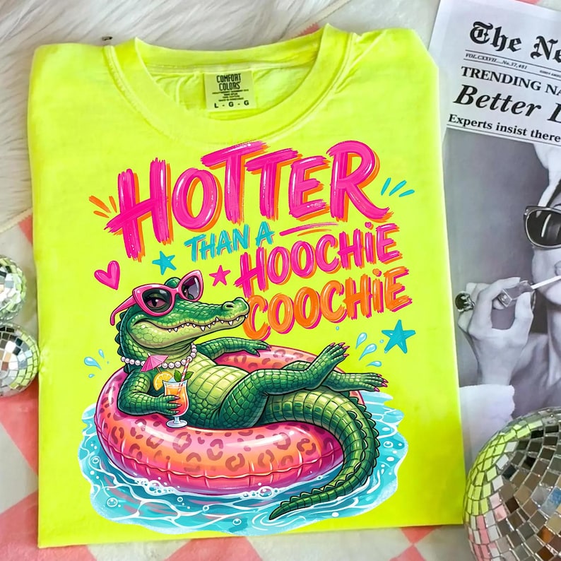 K&ouml;nnte beinhalten: Ein neongelbes T-Shirt mit dem Schriftzug "Hotter Than a Hoochie Coochie" in Pink und Orange. Die Grafik zeigt ein gr&uuml;nes Alligator mit Sonnenbrille und Halskette, der auf einem aufblasbaren Ring schwimmt.