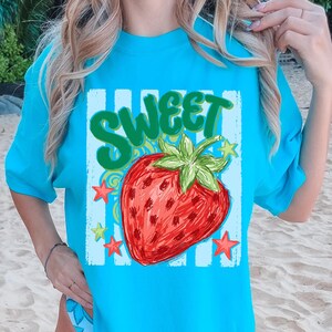 May include: A turquoise t-shirt featuring a large, illustrated strawberry with the word "SWEET" in green above it. The strawberry is red with green leaves, surrounded by stars and a blue and white striped background.