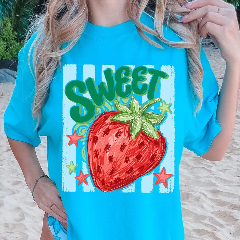 May include: A turquoise t-shirt featuring a large, illustrated strawberry with the word "SWEET" in green above it. The strawberry is red with green leaves, surrounded by stars and a blue and white striped background.