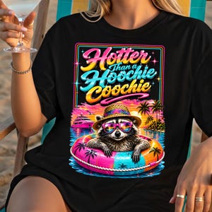 May include: Black t-shirt with a colorful graphic of a raccoon wearing sunglasses and a straw hat, floating in a pink and blue inner tube. The text reads "Hotter Than a Hoochie Coochie". Palm trees and a boat are in the background.