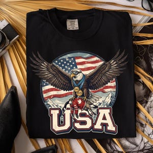 May include: Black t-shirt featuring a graphic of an eagle with outstretched wings, wearing a medal and sunglasses, set against a backdrop of the American flag and mountains. The word "USA" is printed below.