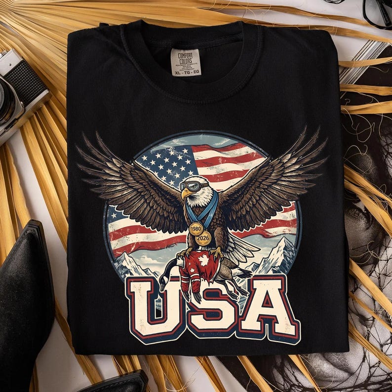 May include: Black t-shirt featuring a graphic of an eagle with outstretched wings, wearing a medal and sunglasses, set against a backdrop of the American flag and mountains. The word "USA" is printed below.