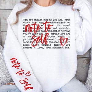 May include: White sweatshirt with red text that reads "You are enough just as you are. Your worth isn't tied to achievements or the opinions of others, it's tossed in your unique light and strength. Even through tough days remember how far you've come and how capable you are of rising again. Speak kindly to yourself. Treat yourself with the same love you'd give someone you care about. Love yourself fiercely, you deserve it. Love, Your Strongest self." The text "note to self" is written in red on the sleeve of the sweatshirt.