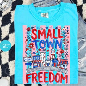 May include: A lagoon blue t-shirt with a graphic design that reads "SMALL TOWN FREEDOM" in red and blue. The design includes illustrations of buildings, flowers, and a water tower, with a pink and white striped background.
