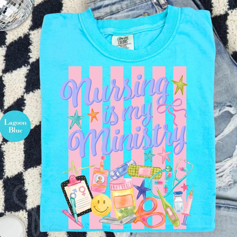 May include: Lagoon blue t-shirt with the text "Nursing is my Ministry" in purple script over pink stripes. The shirt features medical-themed graphics, including a clipboard, pills, and a stethoscope.
