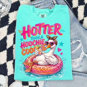 K&ouml;nnte beinhalten: T&uuml;rkisfarbenes T-Shirt mit einer Grafik einer Gans in einem Leoparden-Schwimmring, die eine herzf&ouml;rmige Sonnenbrille und ein Leoparden-Stirnband tr&auml;gt. Der Text lautet "Hotter Than a Hoochie Coochie."