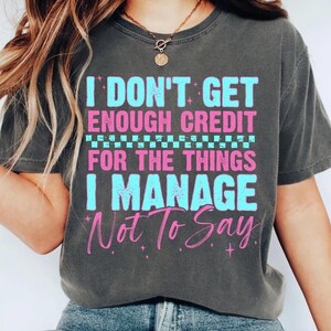 May include: Dark gray t-shirt with the text "I DON'T GET ENOUGH CREDIT FOR THE THINGS I MANAGE NOT TO SAY" in blue and pink. The shirt has a crew neck and short sleeves.