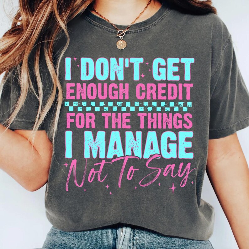 May include: Dark gray t-shirt with the text "I DON'T GET ENOUGH CREDIT FOR THE THINGS I MANAGE NOT TO SAY" in blue and pink. The shirt has a crew neck and short sleeves.