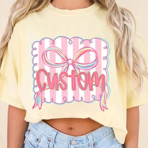 May include: A pale yellow t-shirt with a pink and white striped design. The design features a pink bow and the word "Custom" in pink script. The shirt is a comfortable, casual style.