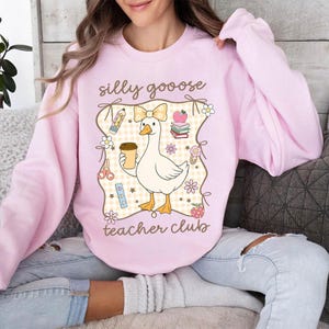 May include: A light pink sweatshirt with a whimsical graphic. The graphic features a white goose holding a coffee cup, surrounded by school-themed illustrations and the text "silly gooose teacher club."