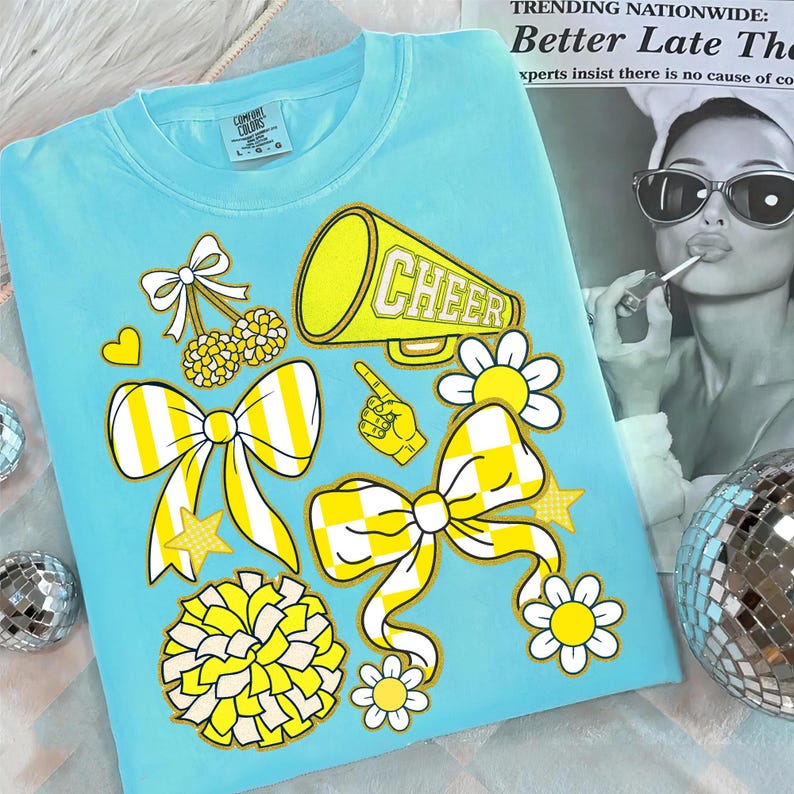 May include: A light blue Comfort Colors t-shirt featuring yellow and white graphics. The design includes a megaphone with the word "CHEER", bows, flowers, and a pom-pom. The shirt has a crew neck and short sleeves.