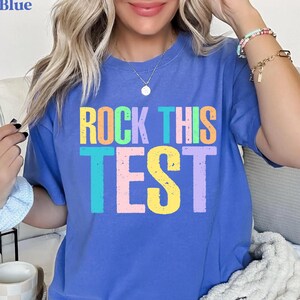 May include: Blue t-shirt with the text "ROCK THIS TEST" in colorful block letters. The shirt is a solid blue color and has a crew neck. The text is in a variety of colors, including yellow, orange, green, purple, and pink.