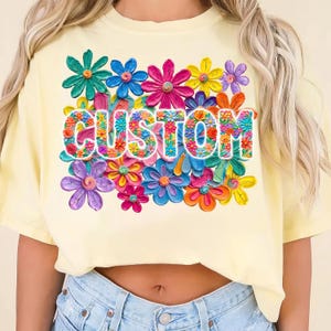 May include: A pale yellow t-shirt featuring a vibrant floral design. The word "CUSTOM" is displayed in a floral font, encircled by an array of flowers in pink, blue, yellow, and purple hues.
