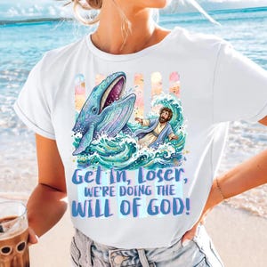 May include: White t-shirt featuring a whimsical illustration of a whale and a figure in water, with the text "Get In, Loser, We're Doing The Will Of God!" in blue and purple. The design has a cartoonish style with a beach background.