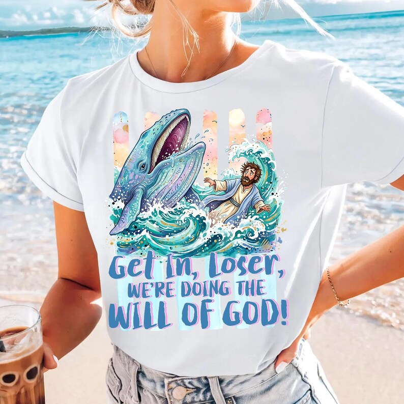 May include: White t-shirt featuring a whimsical illustration of a whale and a figure in water, with the text "Get In, Loser, We're Doing The Will Of God!" in blue and purple. The design has a cartoonish style with a beach background.