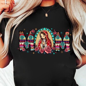 May include: Black t-shirt featuring the word "MOM" in a colorful, patterned design. The "O" is a depiction of a praying figure with a halo, surrounded by pink roses. The shirt has a crew neck.