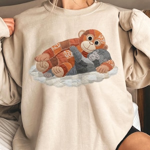 Puede incluir: Sudadera beige con un diseño de aplique de dos monos abrazándose sobre una nube. El mono más grande es de patchwork en tonos naranja y marrón. El mono más pequeño es gris. La nube es blanca y azul claro.
