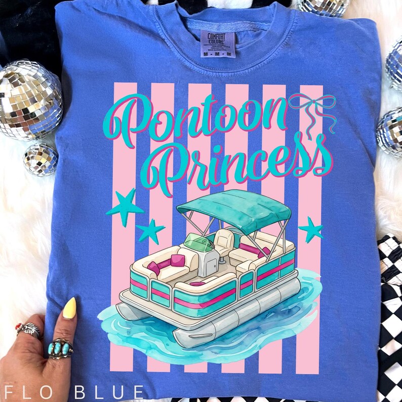 May include: Flo blue Comfort Colors t-shirt with a graphic of a pontoon boat on water. The shirt features the text "Pontoon Princess" in teal script with pink vertical stripes and teal stars. The shirt is a relaxed fit.