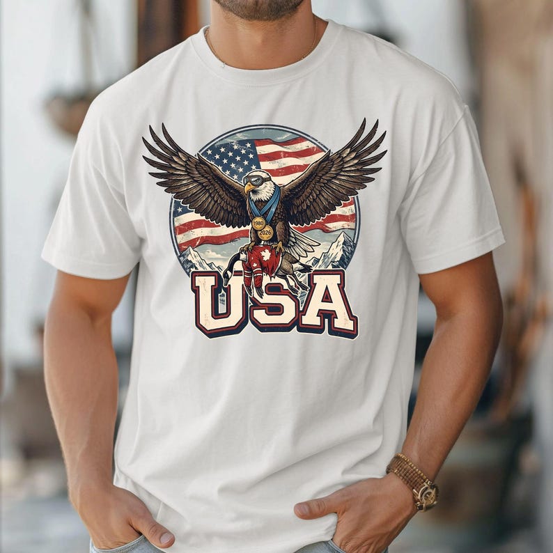 May include: White t-shirt featuring a graphic of an eagle with outstretched wings, a USA flag, and the word "USA" in a bold font. The eagle is wearing medals, and the background includes mountains.