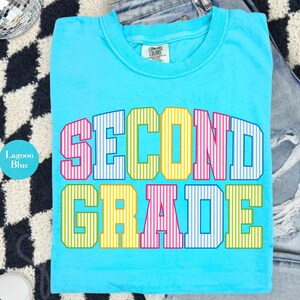 May include: A lagoon blue short-sleeve t-shirt featuring the text SECOND GRADE in a multi-colored, vertical-striped block font. The letters alternate between pink, blue, yellow, and green. The shirt has a crew neckline and a Comfort Colors label.