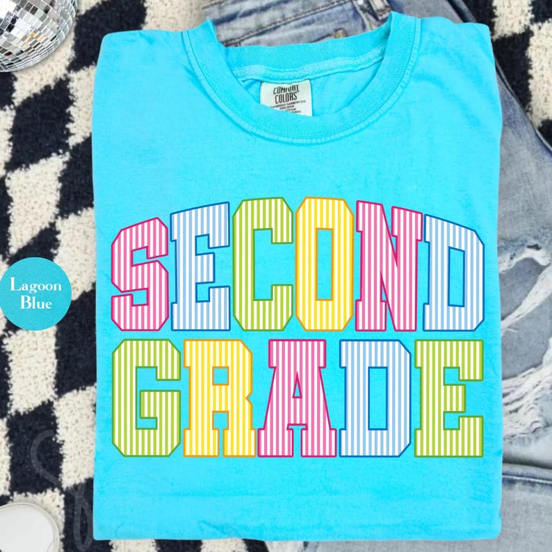 May include: A lagoon blue short-sleeve t-shirt featuring the text SECOND GRADE in a multi-colored, vertical-striped block font. The letters alternate between pink, blue, yellow, and green. The shirt has a crew neckline and a Comfort Colors label.