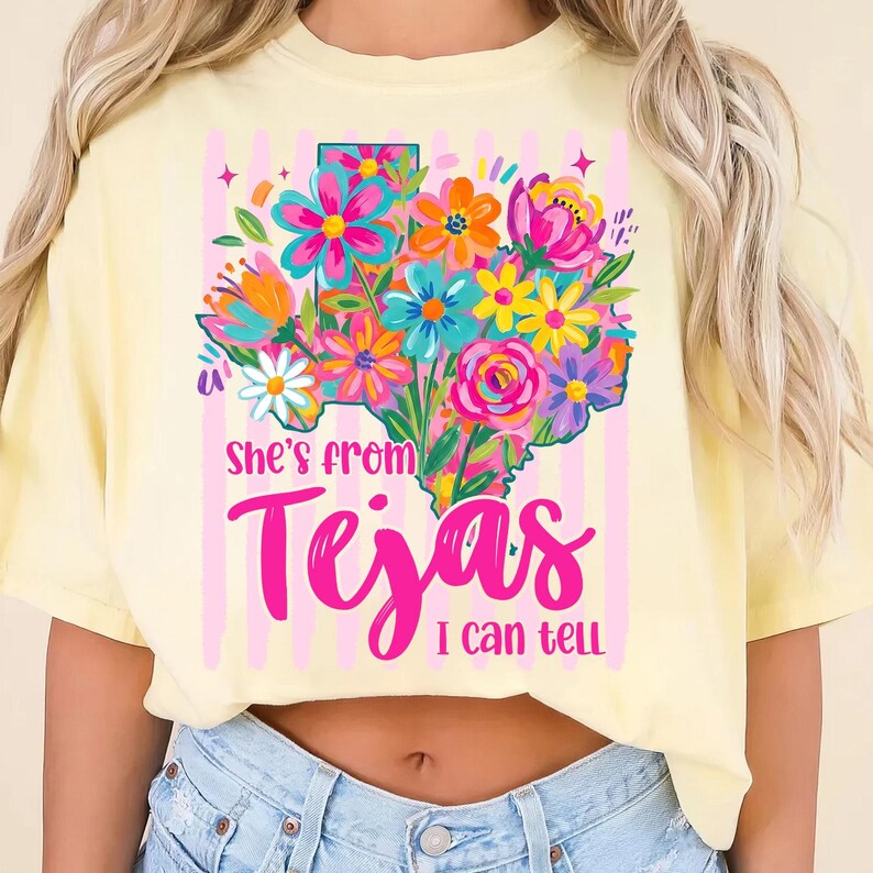 May include: A light yellow t-shirt featuring a colorful floral design in the shape of the state of Texas. The design includes the text "she's from Tejas I can tell" in a vibrant pink script. The background has pink vertical stripes.