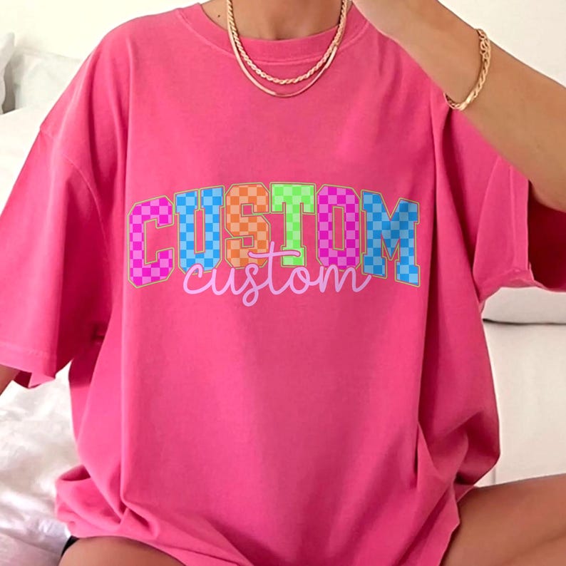 May include: A bright pink t-shirt with the word "CUSTOM" in a colorful, checkered design across the front. The word "custom" is written in a cursive font below the main text. The shirt has short sleeves and a relaxed fit.