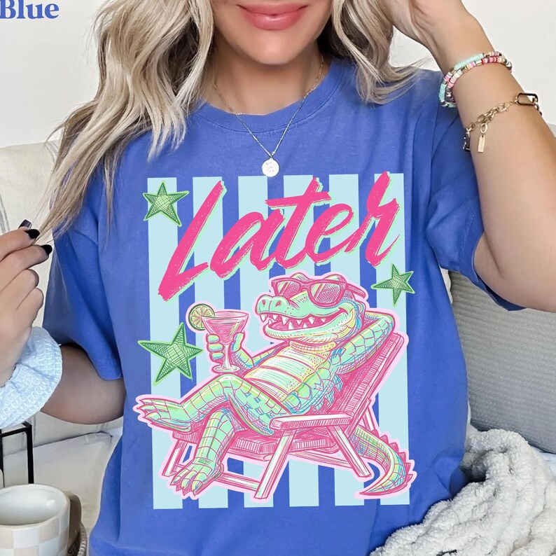 Pode incluir: Camiseta azul com um gr&aacute;fico de um jacar&eacute; relaxado usando &oacute;culos de sol e um coquetel, com a palavra "Later" em rosa. O design inclui listras azuis claras e estrelas verdes.