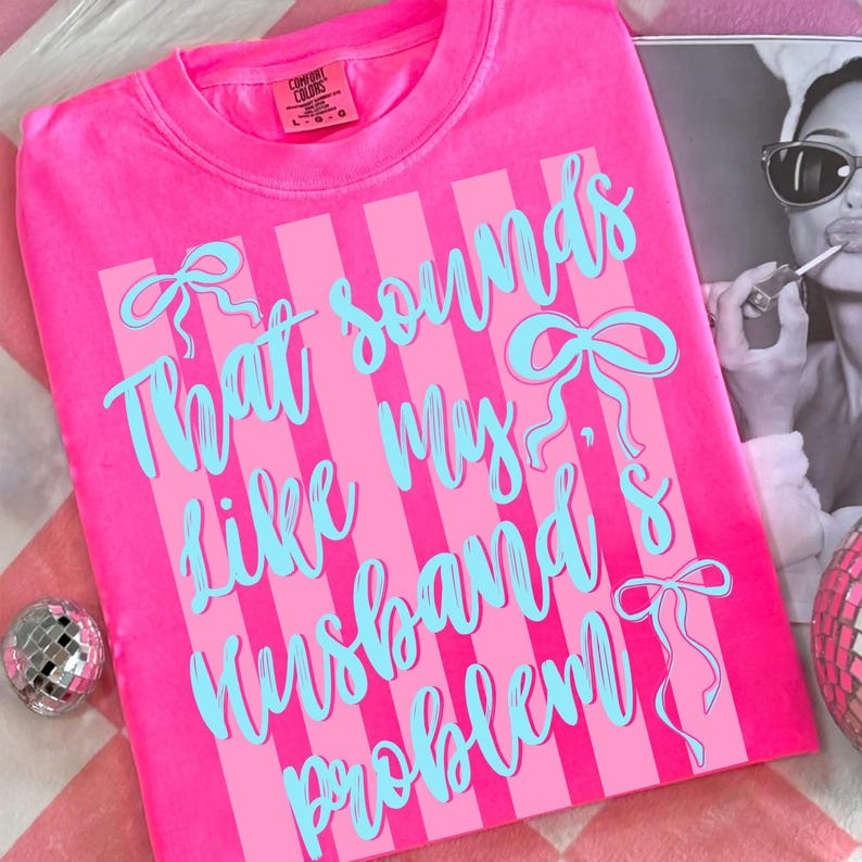 May include: A bright pink t-shirt with the text "That sounds like my husband's problem" in light blue script. The shirt has vertical pink stripes and bow accents. The Comfort Colors brand tag is visible.