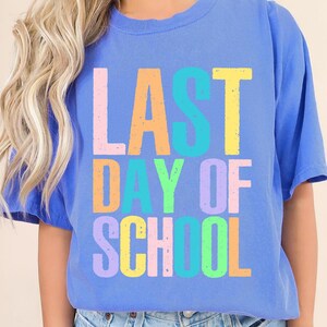 May include: A blue t-shirt with the words "LAST DAY OF SCHOOL" in colorful block letters. The text is in various pastel colors, including pink, orange, yellow, green, and purple. The shirt has short sleeves and a crew neck.