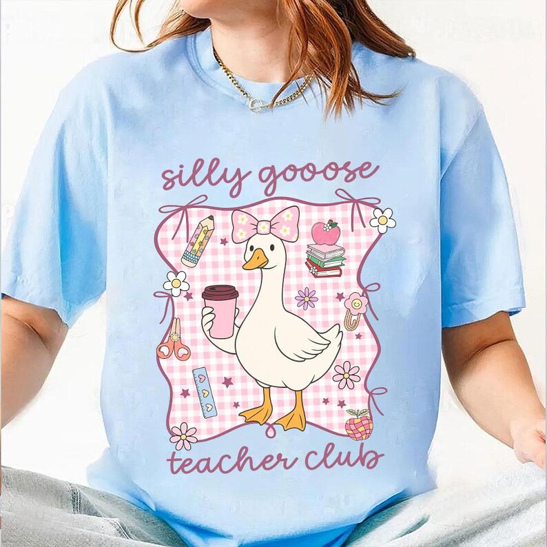 May include: Light blue t-shirt with a graphic featuring a cartoon goose holding a coffee cup, surrounded by school-themed illustrations. The text "silly goooose teacher club" is displayed in a playful font, perfect for teachers.