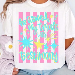 May include: White t-shirt with a pink striped background and the text "He turned my trials into testimony" in a colorful, whimsical font. The design includes star and burst graphics in blue and yellow.