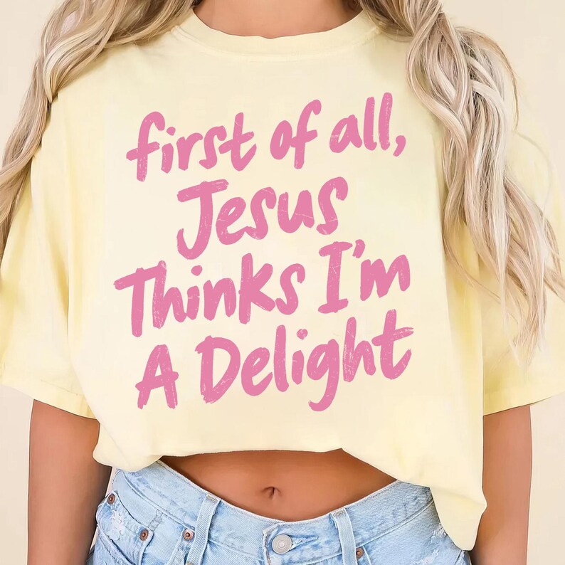 May include: A light yellow t-shirt with the text "first of all, Jesus Thinks I'm A Delight" in pink lettering. The shirt is made of a soft material and has a relaxed fit.