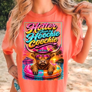 May include: Coral-colored t-shirt featuring a graphic of a Highland cow wearing sunglasses and a pink cowboy hat, floating in a colorful inflatable ring. The graphic includes the text "Hotter Than a Hoochie Coochie" in a vibrant, retro font.