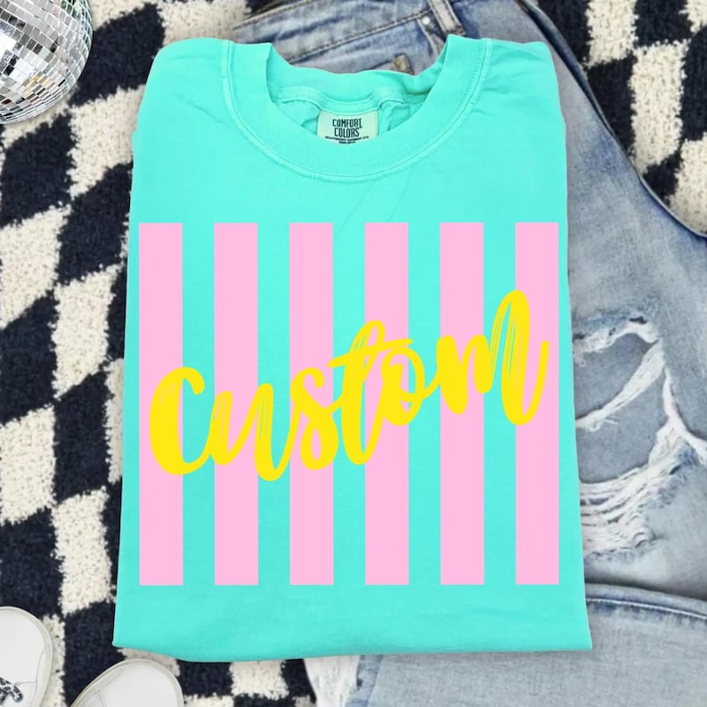 K&ouml;nnte beinhalten: T&uuml;rkisfarbenes T-Shirt mit rosa vertikalen Streifen und dem Wort "custom" in gelber Schrift. Das T-Shirt liegt gefaltet auf einer blauen Jeans. Die Marke "Comfort Colors" ist sichtbar.