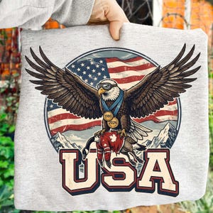 May include: A light gray sweatshirt featuring a graphic of an American bald eagle with outstretched wings, wearing goggles and a medal. The eagle is set against a backdrop of the American flag and mountains, with the word "USA" in bold letters below.