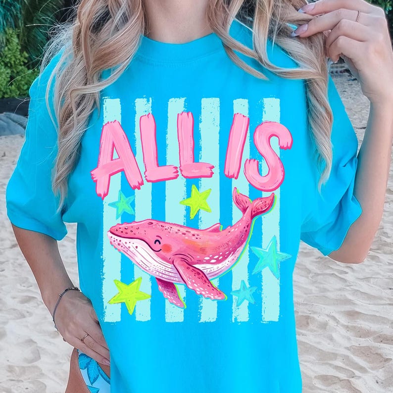 以下が含まれることがあります： ピンクのブロック文字で「ALLIS」という名前が書かれた、ピンクのクジラのイラストが描かれたターコイズ色のTシャツ。デザインには青と黄色の星と縦縞が含まれています。