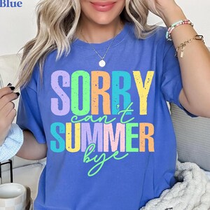 May include: A blue t-shirt with the text "SORRY can't SUMMER bye" in a colorful, distressed font. The text is in shades of purple, green, yellow, and orange. The shirt is a solid blue color.