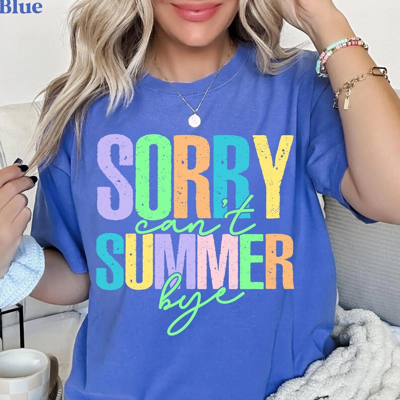 May include: A blue t-shirt with the text "SORRY can't SUMMER bye" in a colorful, distressed font. The text is in shades of purple, green, yellow, and orange. The shirt is a solid blue color.