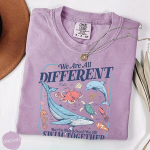 May include: A lavender Comfort Colors t-shirt with the text "We Are All Different" and various sea creatures. The design includes a whale, octopus, dolphin, and other marine life. A silver necklace with a pendant is on the shirt.