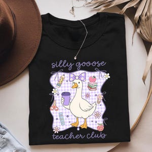 May include: Black t-shirt featuring a cartoon goose holding a purple coffee cup, with the text "silly goooose teacher club." The design includes a purple bow, pencils, books, and flowers. The goose is standing on a purple and white checkered background.